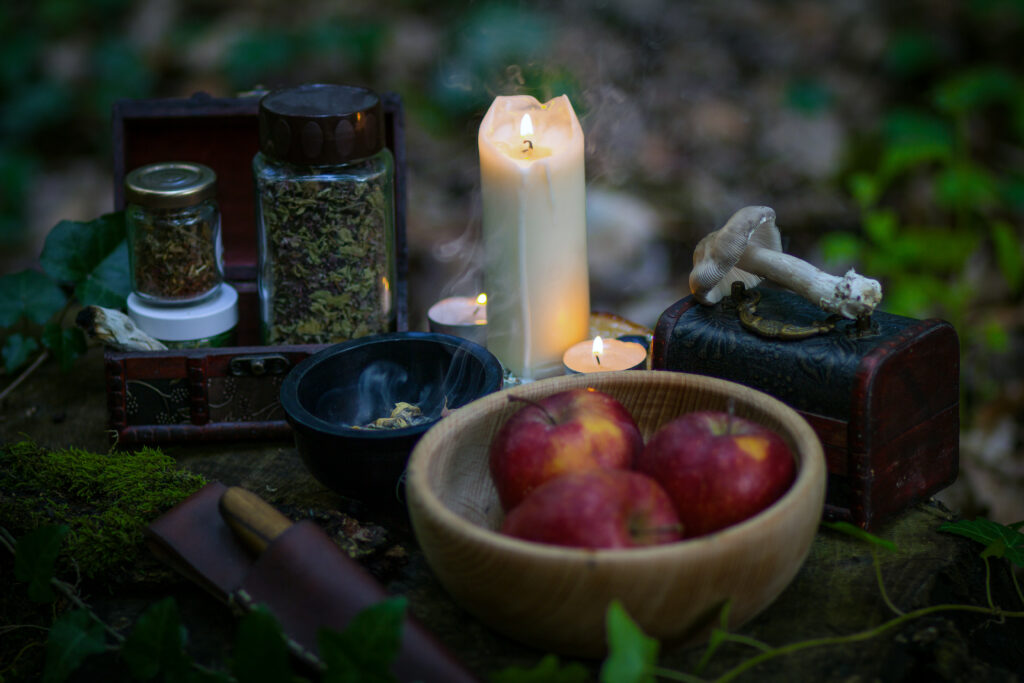 a vida após a morte na wicca. imagem traz elementos de um ritual wicca. vê-se uma vela, cogumelo, um pequeno baú, maçãs e ervas. o cenário é um bosque